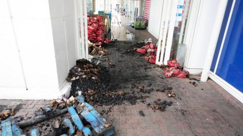 A large amount of burned charcoal and a damage blue pallet in the entrance of a supermarket