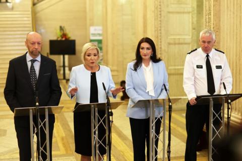Four people stand behind lecterns with microphones, addressing a press briefing in a large, ornate indoor hall with marble walls and tall columns. All are dressed in formal or professional attire, and one person on the right wears a uniform with a visible police badge.
