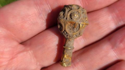 The small, gold object in the palm of someone's hand. It is covered in dirt but the bright gold can be seen underneath it. It has a stem and round top with circular patterns carved into it.