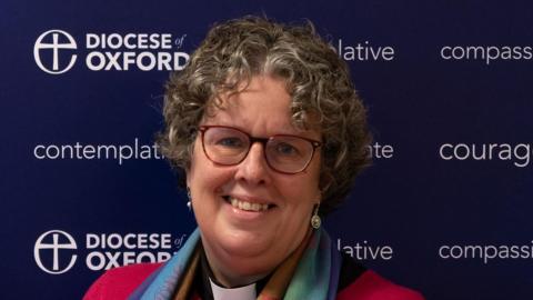 Reverend Canon Mary Gregory has brown curly hair and wears red-trimmed glasses. She has a black shirt and white dog collar on, with a colourful scarf and pink cardigan.