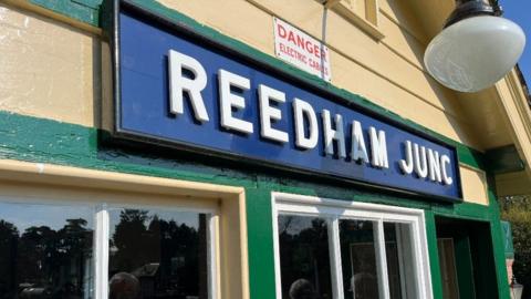 Close up of the yellow and green signalling box with a blue sign with white writing that says Reedham Junction