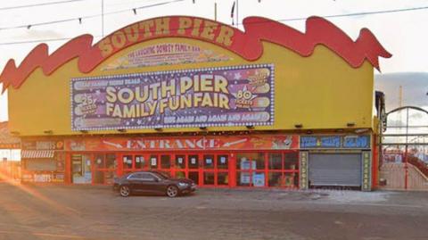 The entrance to South Pier, a large rectangular building painted yellow and red. It has a huge sign saying South Pier in yellow under a red roof shaped like a wave and many large red glass entrance doors with a small shop either side