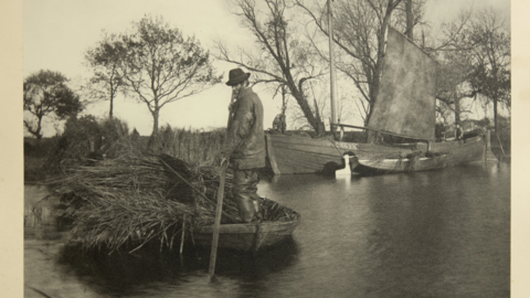 Black and white image of a man pushing through a river on a small boat loaded with reeds with a small wherry boat in the background
