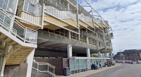 A bus shelter in Sherborne Road in Newton Abbot. It shows a multi-storey car park with a stairway on the side of the building. Below is the us shelter with people waiting for a bus.
