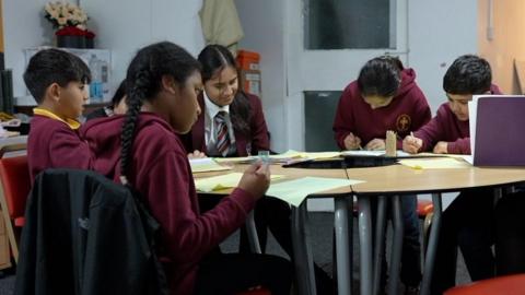 A group of six male and female school pupils of varying ages sitting around a table writing in notebooks and on worksheets. They are all wearing maroon school uniforms.