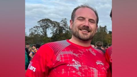 A man smiling directly at the camera. He is wearing a red patterned rugby top. There are trees in the background.