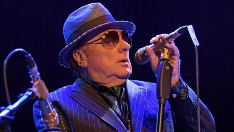 Image of Van Morrison wearing pinstripe navy suit singing into a microphone. He is wearing a bowler hat and tinted glasses.