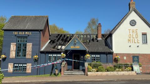 Police tape runs across the entrance to a blue-coloured pub building, with a badly damaged roof caused by a fire in view on a sunny day