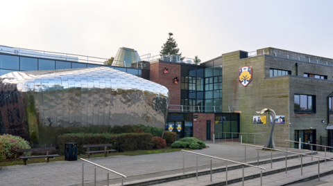 general view of St Regis CE Academy in Wolverhampton - rectangular buildings with a large silver round section at the front near the entrance. A logo can be spotted on the building on the right