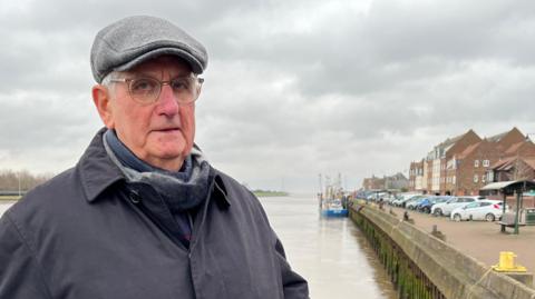 Peter Harvey is wearing a grey flat cap and dark navy coat. He is standing in front of The Wash - a large tidal body of water. A row of cars are also parked on land behind him, next to some buildings.