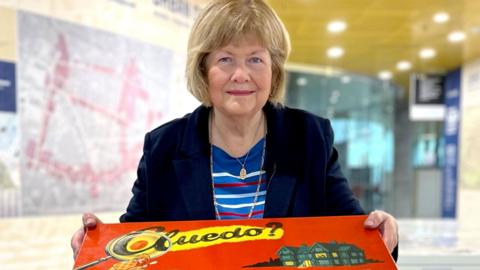 Marcia Lewis is holding one of the original versions of the Cluedo board game in its red box. She is wearing a dark blue suit jacket.