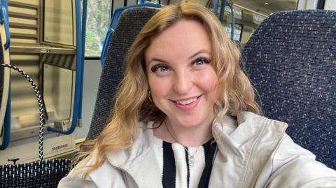 The image shows a woman smiling on a train; she is wearing a white coat and a black and white striped top underneath.
