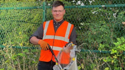 Man wearing glasses and a bright orange vest in front of bushes and woodland.
