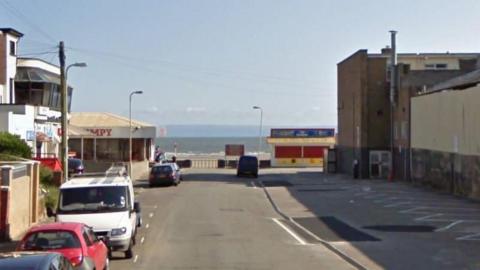 The end of Mackworth Road, leading to the beach and the sea at the end. Vehicles are parked on the left hand side, with empty parking spots to the right. A Wimpy restaurant can be seen in the background. 