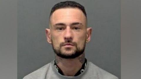 A police custody image of Kieran Humphreys. He has dark hair and a dark beard and is looking straight ahead. He has tattoos on his neck and is wearing a grey top.