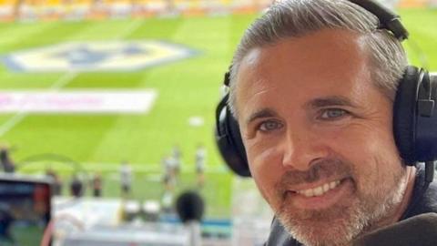 Ryan is in the ground with headphones on ahead of a match. He has grey hair and a beard and us turning around form his desk to have the photo taken. The pitch can be seen in the background.