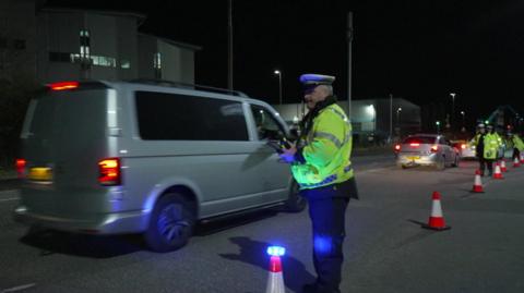 A police officer stands by a row of cones at the ferry port as a cars file past.