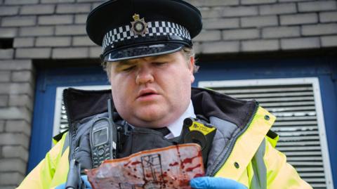 Ethan Lawrence as police officer Ben in Boat Story. Ben wears a police hat and high-vis coat with a walkie talkie and camera attached to his vest. He holds a bloodied piece of paper in his blue-gloved hands 