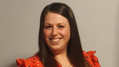 Abby Storey  is smiling brightly at the camera. She has long brown hair and is wearing a spotty red dress.