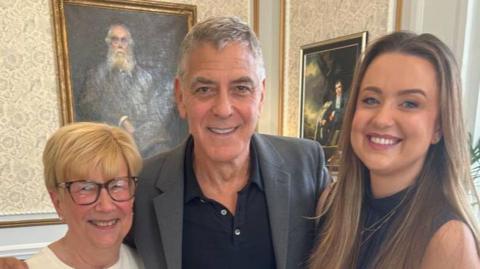 Three people, two women and George Clooney, posing together indoors on a patterned carpet, standing in front of framed artwork, dressed smartly with one person resting an arm around another.