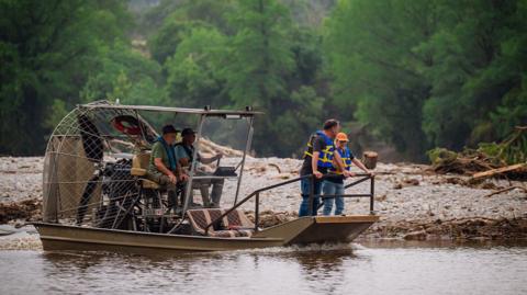 Four people in a boat on a river with life vests on. There is a lot of debris in the background.
