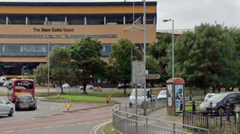 A 1980s phone box with tatty posters visible on two sides stands on the edge of an urban roundabout flanked by a grass verge and lamp posts. In the background, the stand of a football stadium is visible in yellow brick, with a black sign reading The Stan Cullis Stand.