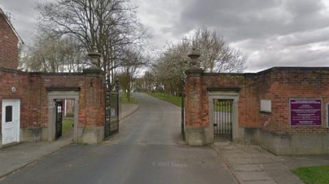The main road entrance into Herringthorpe cemetery.  There is a tall red brick wall around the perimetre.  The road goes through a gateway with black iron gates which are opened.  There are pedestrian gates on wither side, to the left the gate is open, the pedestrian gate to the right is closed.