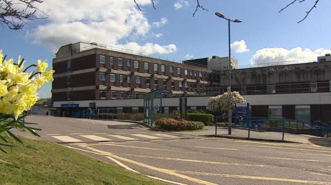 The outside of a hospital on a clear sunny day. The building has lots of windows and has the usual NHS branding on the front of it. There is a road in front of it with some flowers to the left of the pictures. 