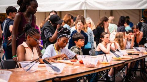 A group of children sat on black chairs behind a long wooden table. On the table are plastic bowls, rolling pins and playdough. Groups of children and teenagers are stood gathered behind the table.