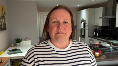 Julie looking directly down the camera. She is standing inside, with a kitchen area behind her. She is wearing a top with horizontal white and black stripes. She has light brown, shoulder-length hair and blue eyes.  