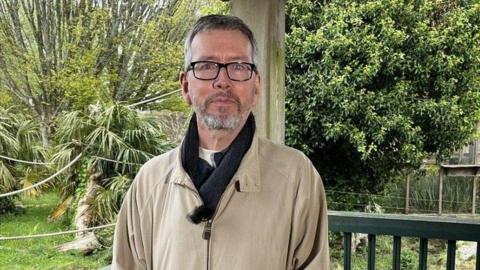 A picture of Matthew Hatchwell wearing a cream coat. Behind him is trees and part of the zoo.