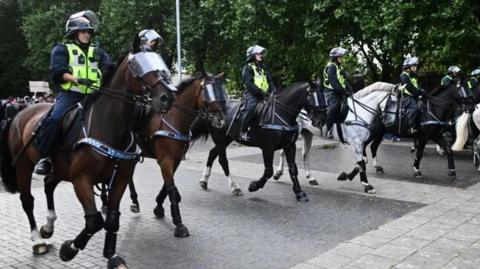 Seven police officers with horses in Bristol. The officers are all wearing helmets and high vis jackets over their uniforms. The horses also have shields over their faces.
