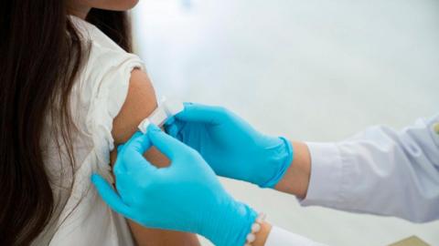 A female child with long brown hair and a white top is getting a white plater stuck onto her arm following an injection. They person putting the plaster on is wearing light blue gloves, a pale blue top and has a pearl bracelet on one of their wrists. 