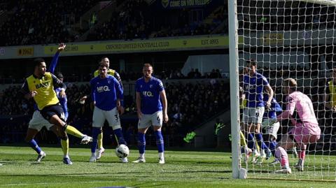 Footballer Myles Peart-Harris strikes a football for Oxford United in a crowded penalty area during a Championship match at home to Watford. The Oxford players are wearing a yellow kit with dark blue shorts and trim details, while Watford are wearing blue shirts and socks with white shorts. A goalkeeper in a predominantly pink kit is visible on the goal line, as is a packed stand full of fans at the Kassam Stadium