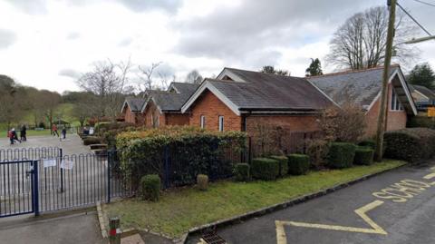 Single story pitched roof brick building with blue metal fence and hedging a playground with children running around is to the left of the building with fields seen beyond.