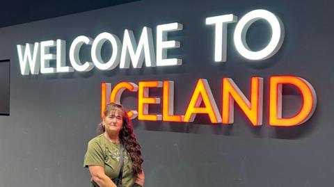 A woman with long brown hair wearing a green t-shirt standing in front of a Welcome To Iceland sign at an airport