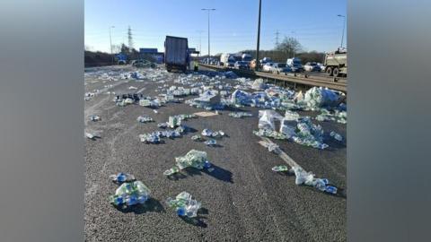 Multiple water bottles in clear and blue packaging are strewn across the motorway. A lorry and police car can be seen in the distance. Traffic travelling in the other direction appears to be queueing.