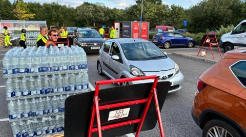 Piles of bottles of water with cars next to them in a car park