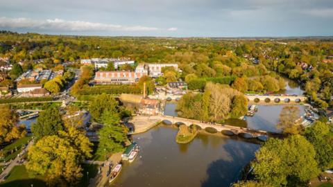 An aerial view of the centre of Stratford-on-Avon with trees on both sides of a wide river and large buildings seen between them. There are two large bridges crossing the river 