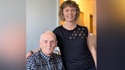 An elderly man in a buttoned up shirt sits next to a younger woman with fair curly hair who stands at his side.
