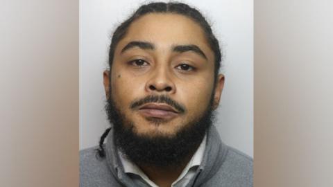 Mugshot of Caleb Fleming, with braided, long black hair, a white shirt and a grey sweatshirt 