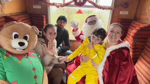 Jessica Lymn with her son Rory is sitting in a carriage with Santa, a woman wearing a Santa cloak and a life-size teddy bear. Father Christmas and Ms Lymn are waving at the camera. They are sitting in an enclosed carriage decorated with tinsel.