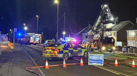 Several emergency service vehicles parked on a road at night with orange cones blocking the street.