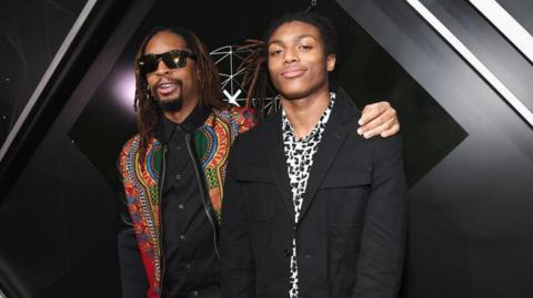 Rapper Lil Jon poses for a photo with his sone Nathan Smith. Jon wears a colourful jacket and black shirt while Smith is dressed in black and white