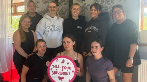 Izzy, who leads the group is sat with a group of women. She's holding a pink circular sign that says the name of the community group 'Yes I Can!' on the front with a drawing of a bow underneath. They appear to have just finished a pilates session, they're wearing workout clothes.