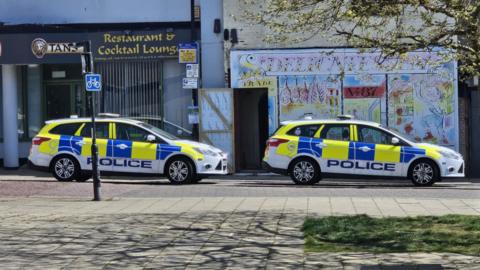 The side-profile of two police cars parked outside shops in Lowestoft town centre in sunny weather. There is a paved area in the foreground. Behind the cars are a restaurant and cocktail lounge and what appears to be a colourful cafe frontage.