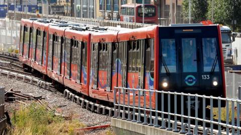 Red DLR train on some track in London, with a road running parallel to it and a red London bus in the background. In the foreground you can see the track and some grass and discarded sections of track and some metal railings.