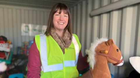 A woman with shoulder length dark brown hair smiles towards the camera. She is wearing a dark pink long sleeve top and a light brown patterned scarf with a yellow and silver high visibility vest on top. She is holding a light brown stuffed hobby horse. The background is blurred but there are piles of donated toys inside a grey shipping container. 