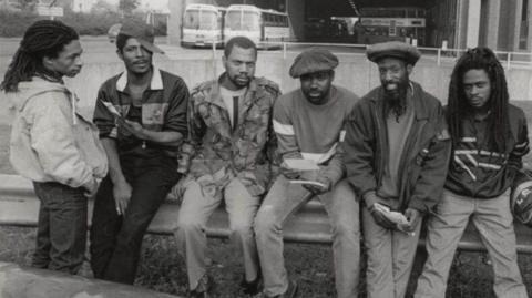 A group of men in front of a bus station, with of them five sitting down and one standing up. The image is black and white. Three of the men are wearing hats.