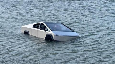 A silver Tesla Cybertruck seen partially submerged in water, which has risen half way up the wheels, in a lake.